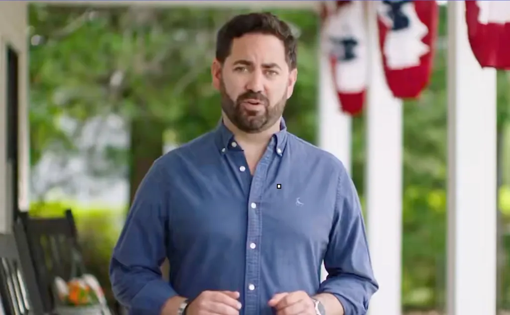Mike Lawler, a man with a beard, wearing a blue button-down shirt, speaks outdoors on a porch.