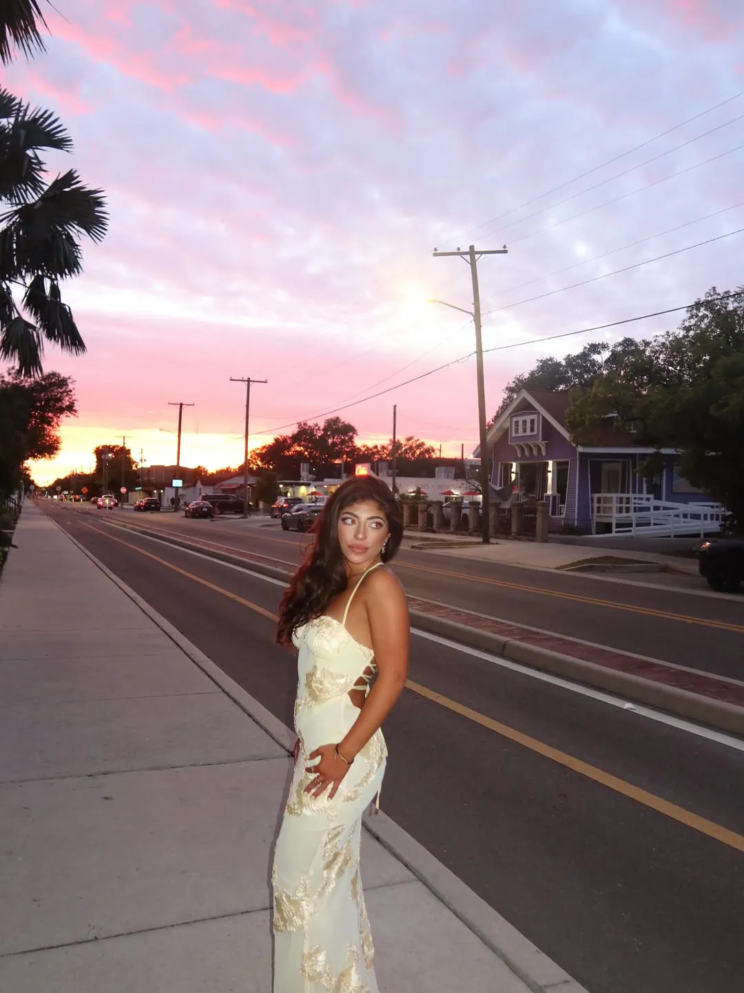 Milania Giudice in a pale yellow dress with gold embroidery stands on a sidewalk against a pink and purple sunset.