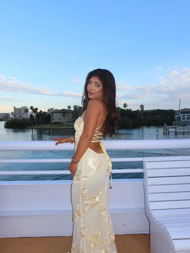 Milania Giudice in a pale yellow dress with floral embroidery, looking over her shoulder from a boat.