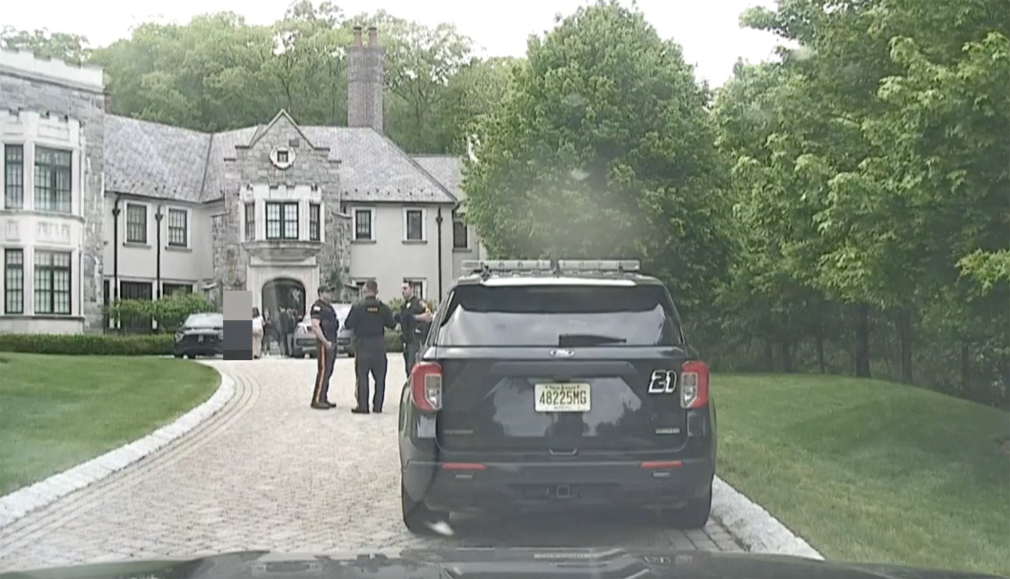 A bodycam view of a police car with flashing lights on the driveway of a large house, with officers in front of the house.
