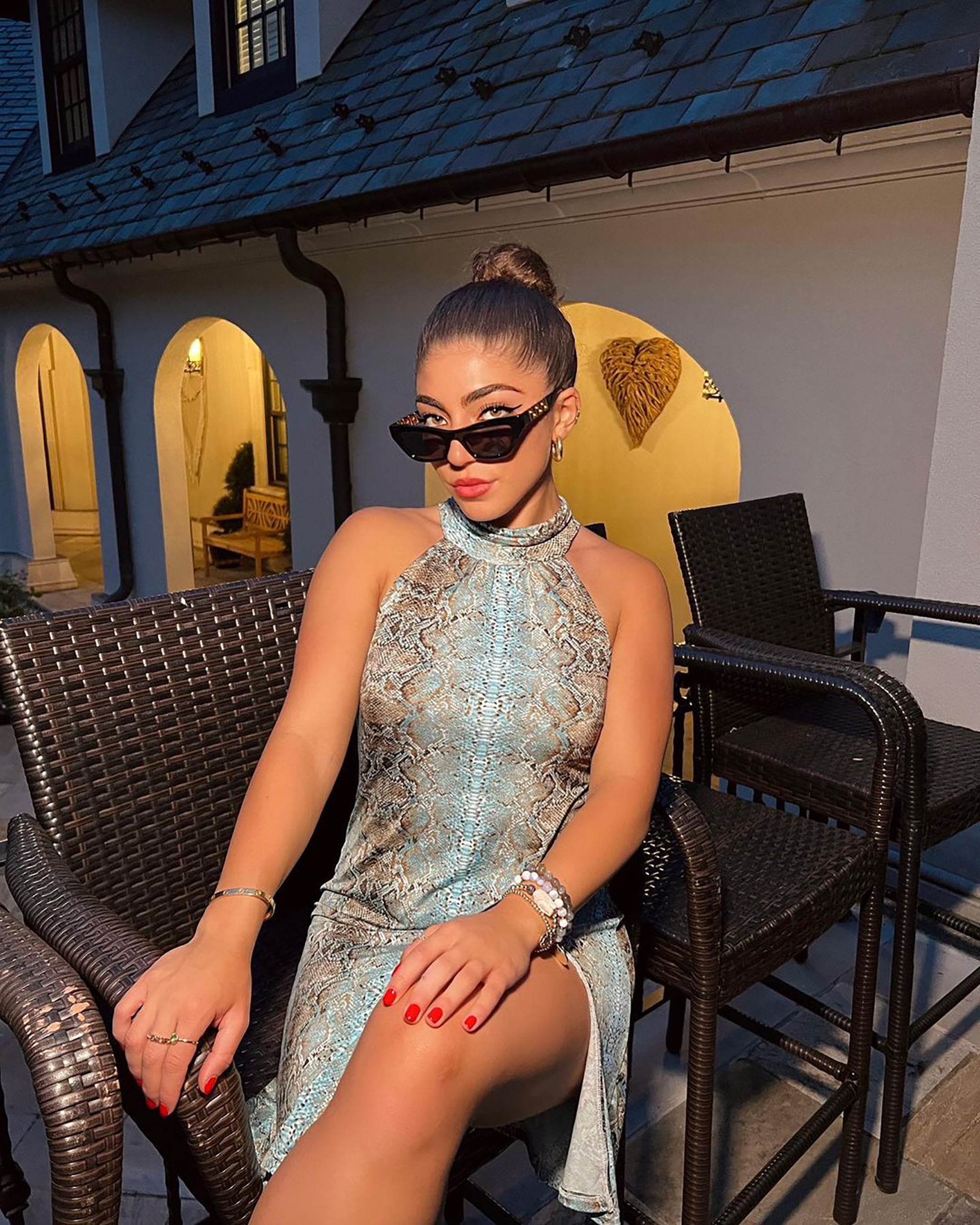 A woman wearing a snakeskin print halter dress and sunglasses sits in a wicker chair.