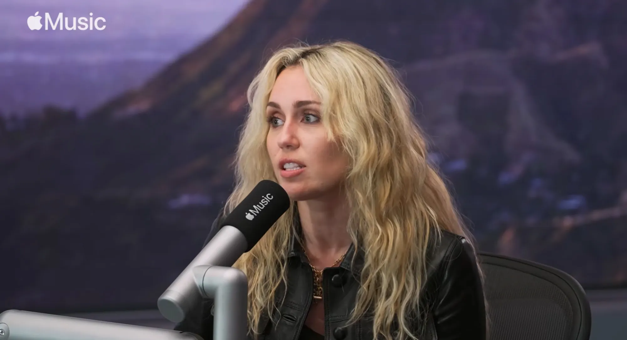 Miley Cyrus with blonde curly hair talking into a black Apple Music microphone.