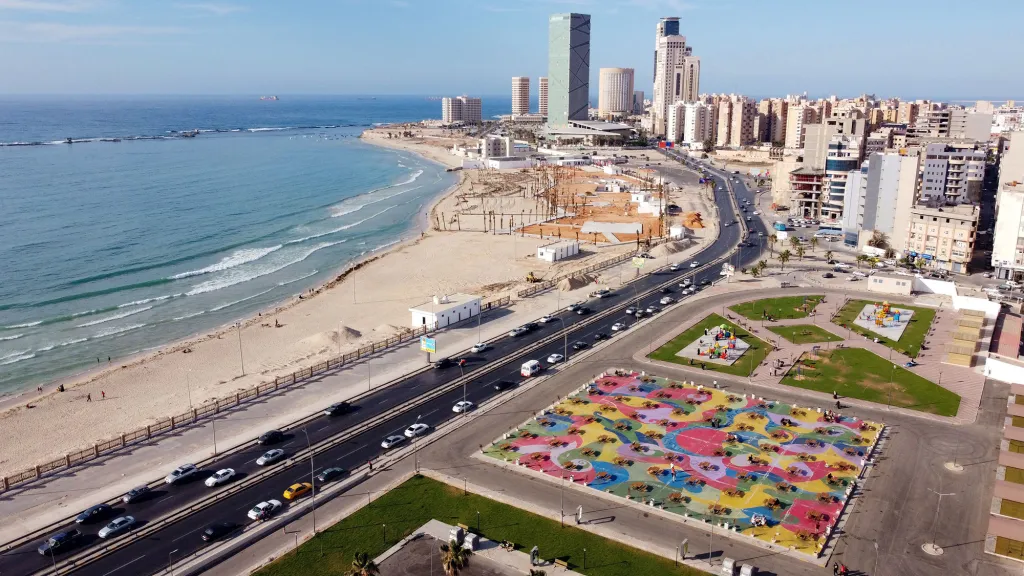 Aerial view of a coastal city with a road, beaches, and buildings, including a colorful recreational area with picnic tables in the foreground.