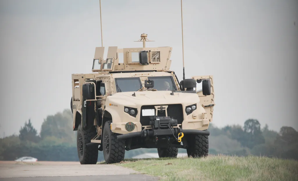 A tan military vehicle with its doors open is parked on a road next to a grassy area.