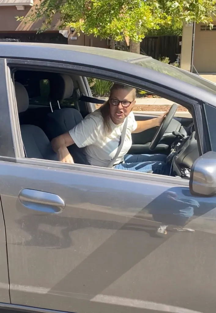 A woman in a gray car yells while looking back and holding the steering wheel.