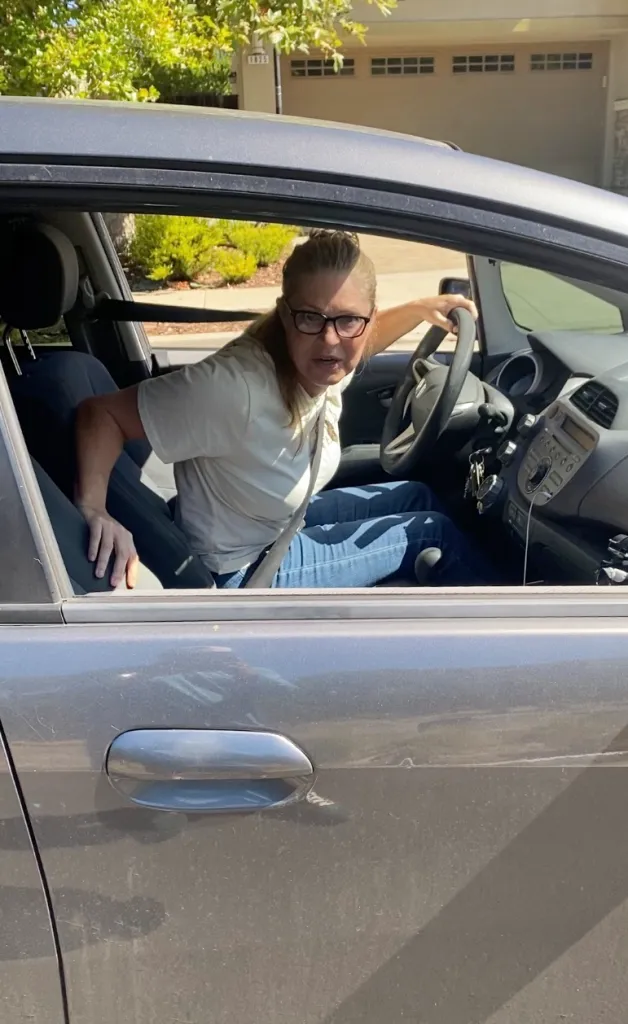 A woman sitting in the driver's seat of a car, leaning forward and looking intently out the open window.