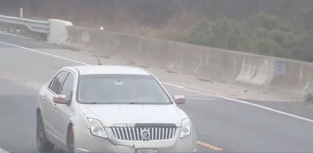 A silver Mercury sedan driving on a highway, with a concrete barrier and some vegetation in the background.