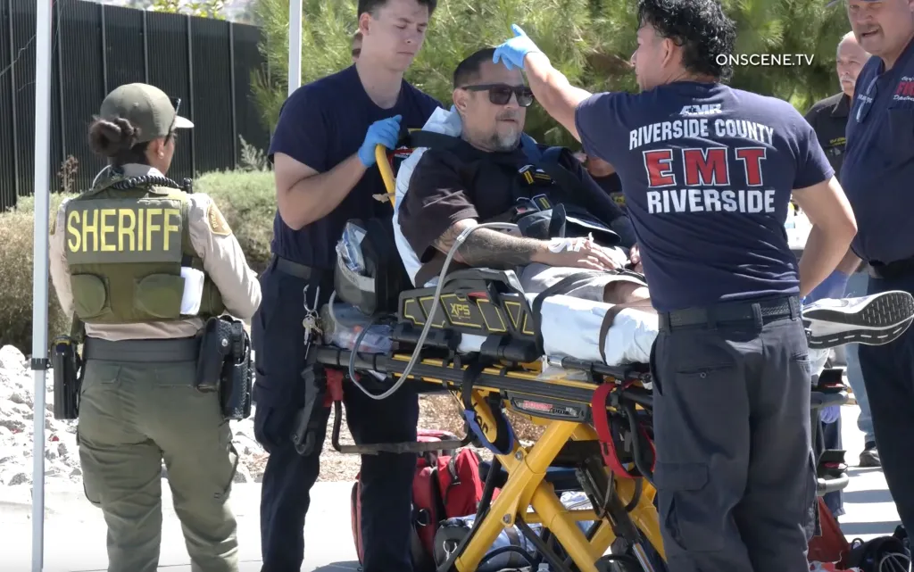 A man in sunglasses and a black shirt is on a gurney, being attended to by three EMTs and observed by a sheriff's deputy.