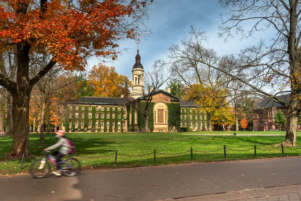 Nassau Hall with its clock tower stands behind a green lawn and trees with autumn foliage, as a cyclist rides by.