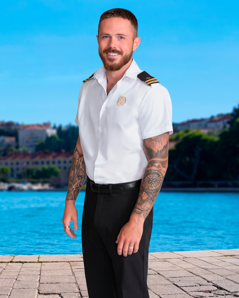 Nathan Gallagher smiling at the camera in a white shirt with captain's epaulets, his arms covered in tattoos, with a background of blue water and distant buildings.