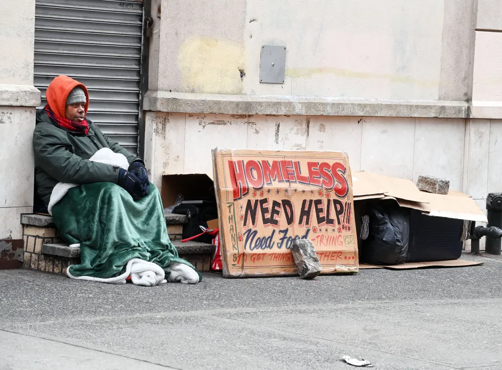 A homeless man, wrapped in a green blanket, sits on the steps next to a