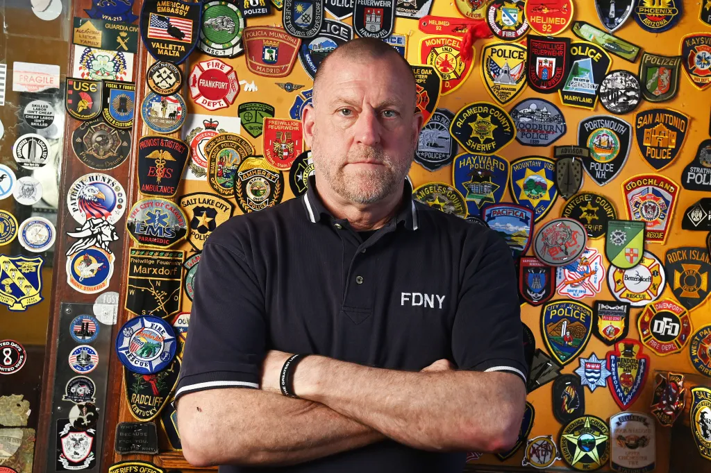 Retired firefighter Tim Brown, wearing an FDNY shirt, stands in O'Hara's Bar & Restaurant in front of a wall covered in over 7,000 first responder patches.