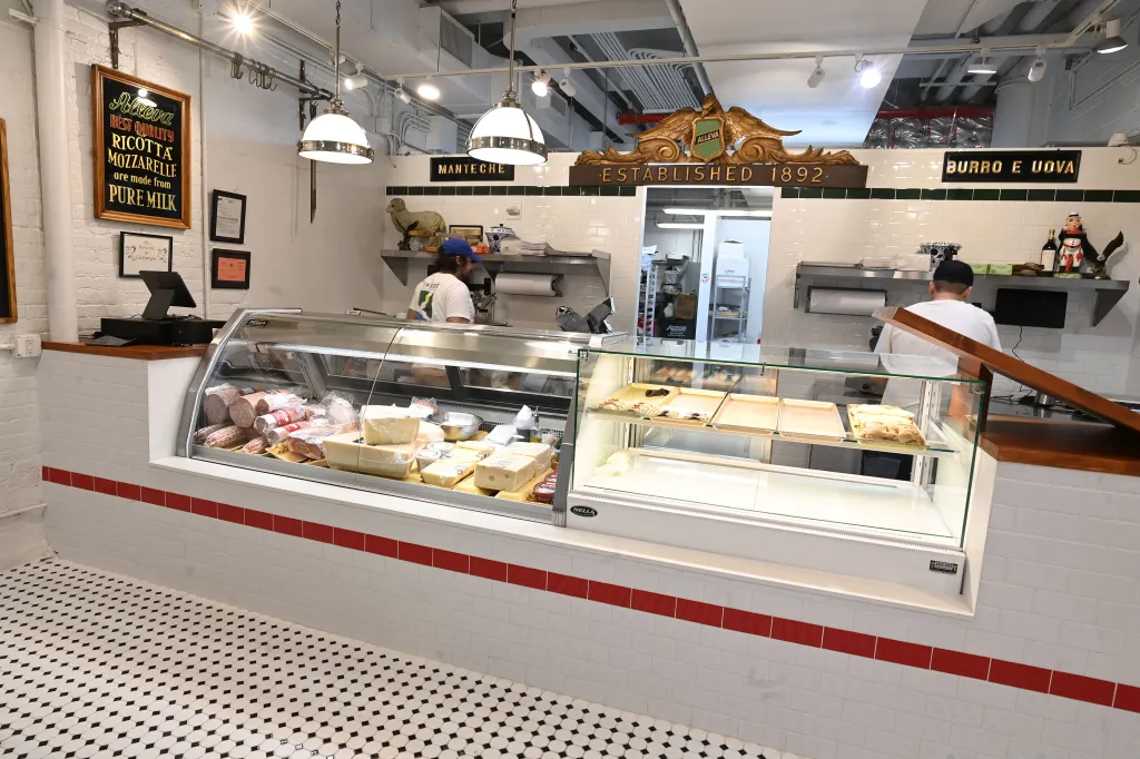 Interior of Alleva Dairy in Chelsea Market with two employees stocking display cases of cheeses, meats, and pastries.