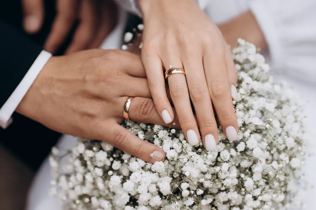 Newlywed couple's hands with wedding rings resting on a white bouquet.