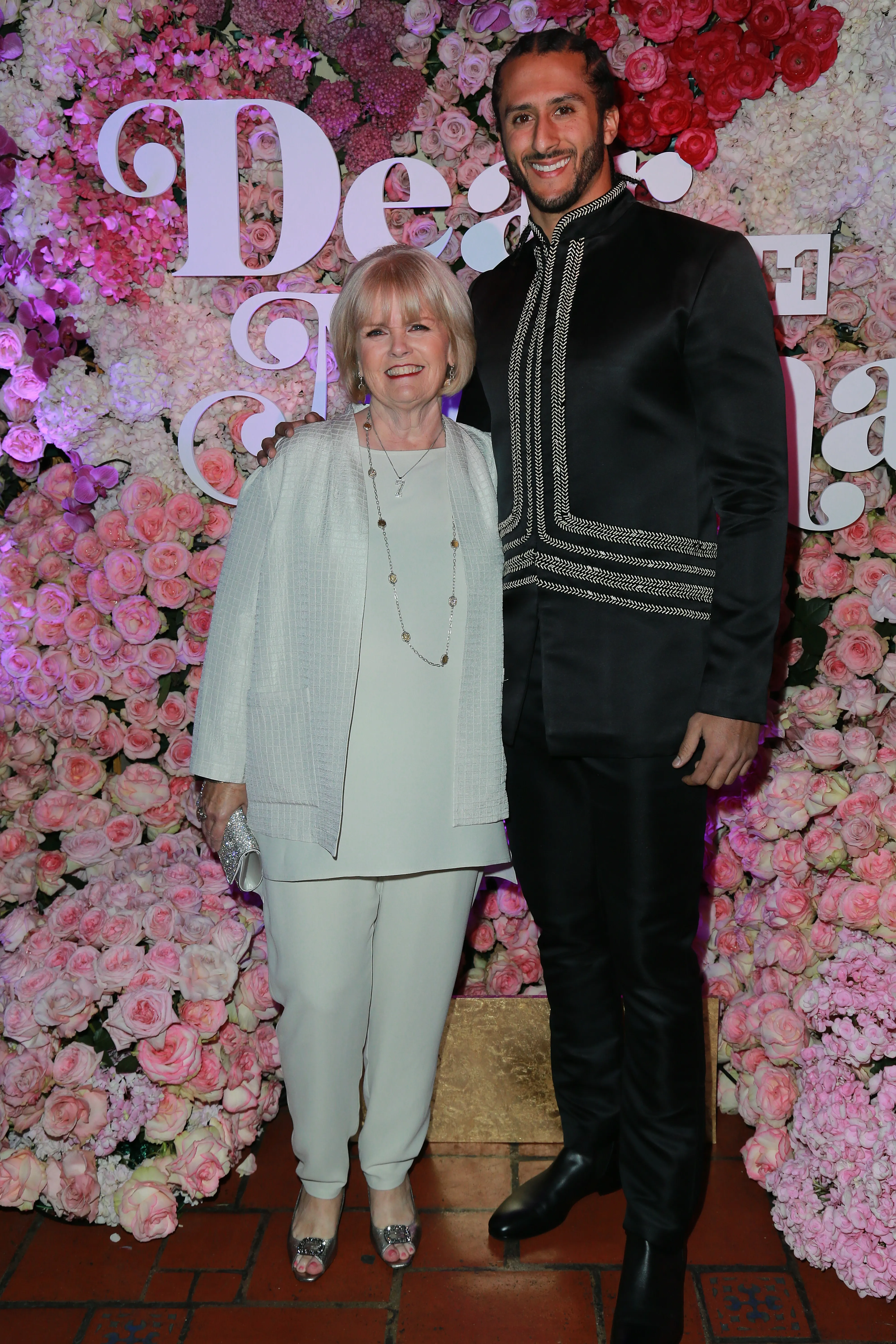 Colin Kaepernick in a black embroidered suit and Teresa Kaepernick in a light pantsuit pose in front of a wall of pink and white roses.