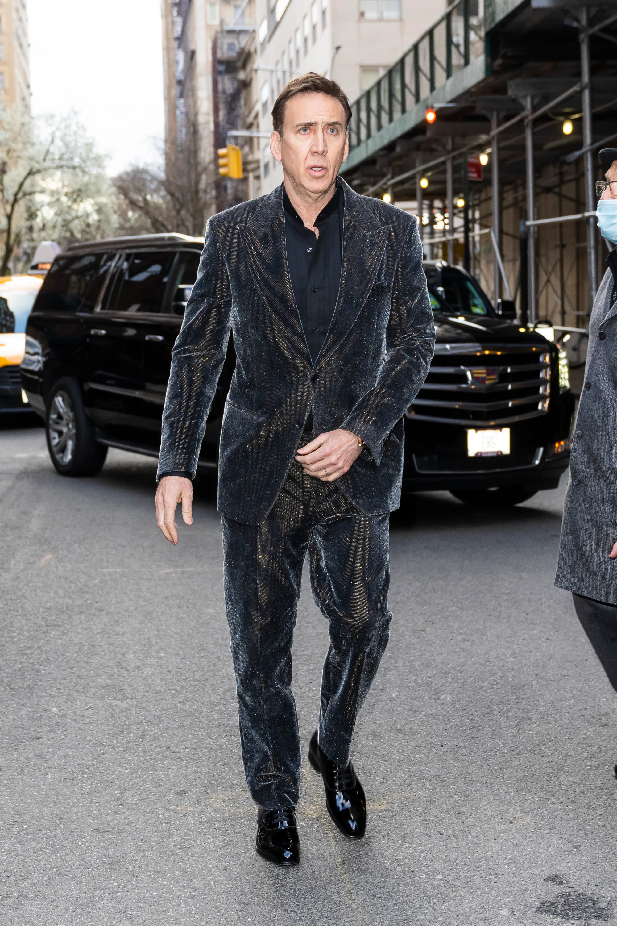 Nicolas Cage walks down a street in a dark, patterned suit.