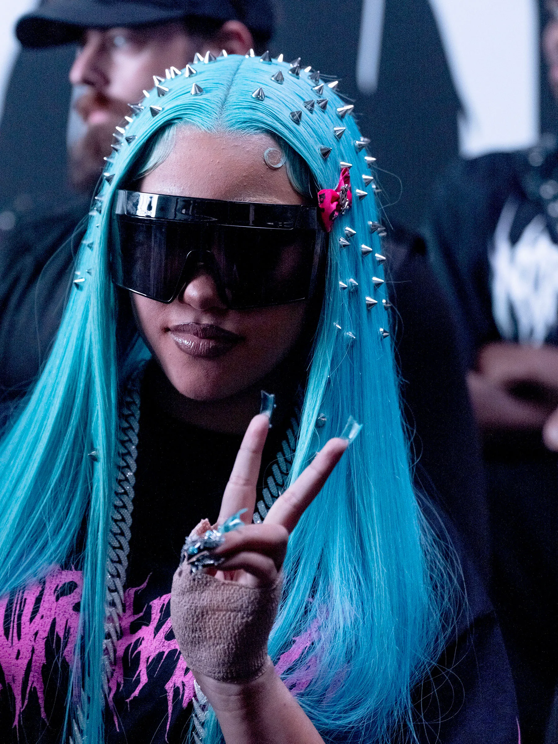 North West wearing blue hair with spikes, large dark sunglasses, and a black shirt with pink lettering, holding up a peace sign.