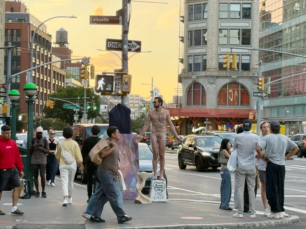 A naked artist named Psuperhamb stands on a soapbox with writing on his body asking for a benefactor, on a New York City street corner.