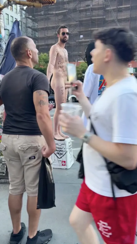 A naked artist standing on a soapbox with writing on his body.