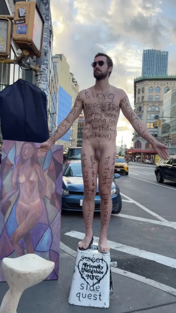 A man with text written across his nude body stands on a street corner next to a painting of a nude woman and a sculpture of a whale tail.
