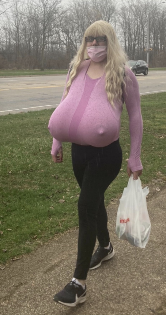 Kayla Lemieux, a teacher, wearing large prosthetic breasts under a pink top, black pants, and a pink mask, walks on a sidewalk while carrying a white plastic bag.