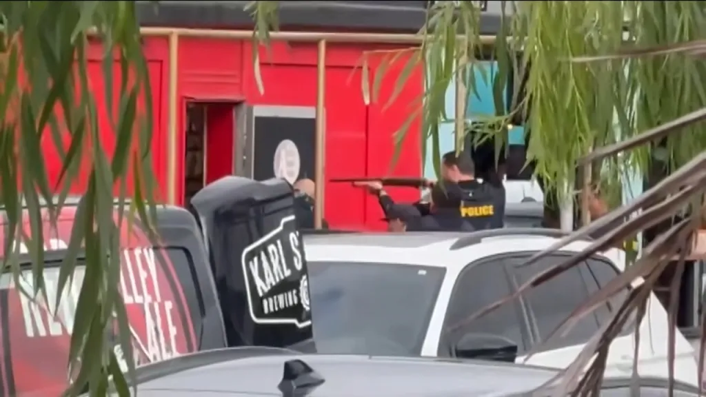 Police officers, one holding a rifle, standing on and next to a white SUV in front of a red building, with foliage in the foreground.