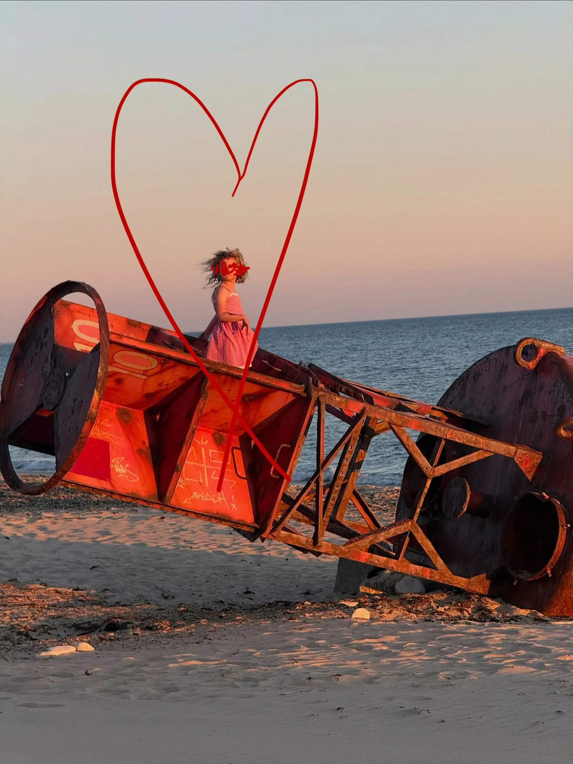 A young girl in a pink striped dress stands on a large, red, rusty buoy on a beach at sunset, with a red heart illustration drawn around her.