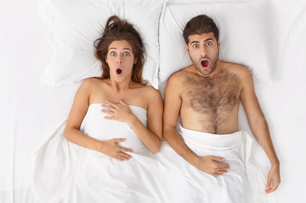 Overhead shot of a shocked woman and man lying in bed under a white sheet, mouths agape.