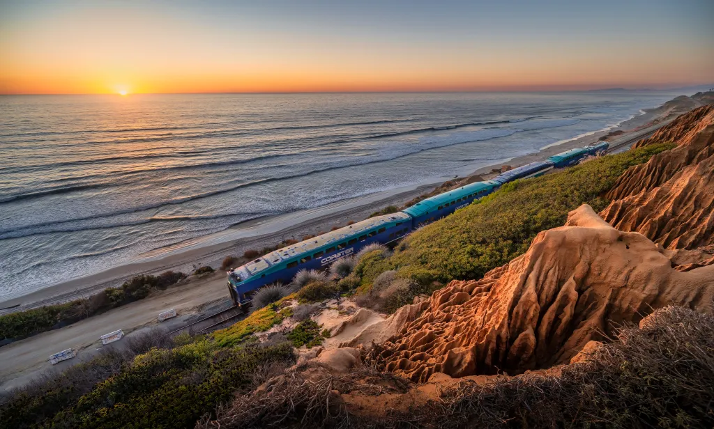 An aerial photo of a blue and green Pacific Surfliner train traveling along a coastline in Del Mar, California, during sunset.