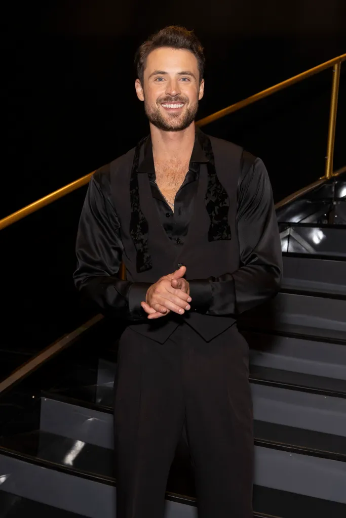 Dylan Efron smiling in a black shirt, vest, and trousers with a gold railing and stairs in the background.
