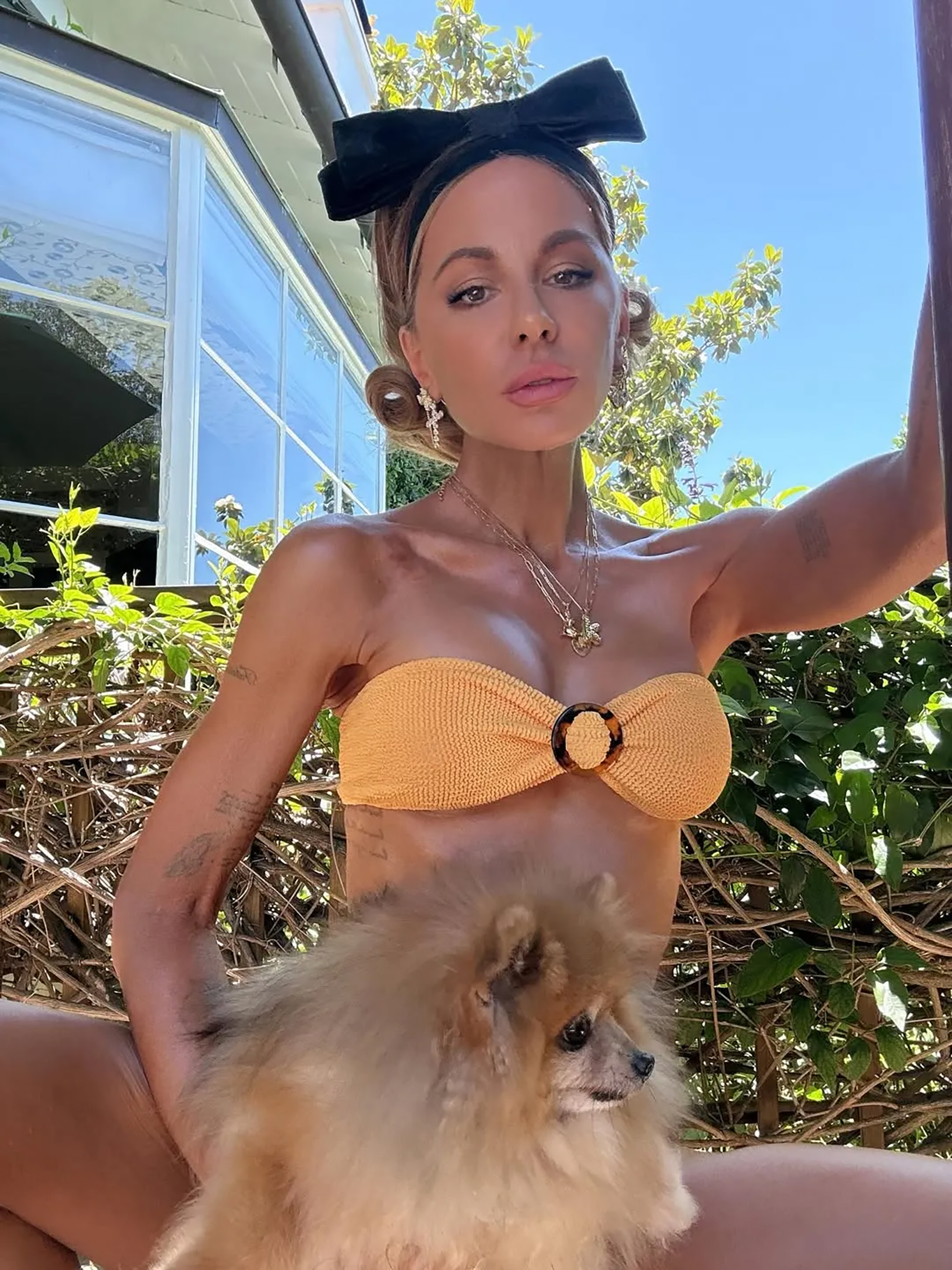 Kate Beckinsale posing outdoors in a yellow bandeau top and black bow headband while holding a fluffy brown dog.