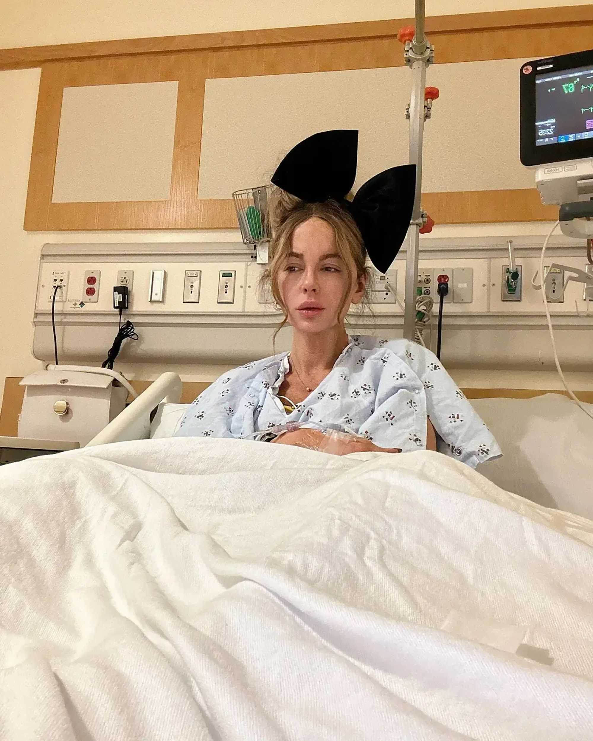 Kate Beckinsale sitting up in a hospital bed with an IV in her arm and wearing a large black bow in her hair.