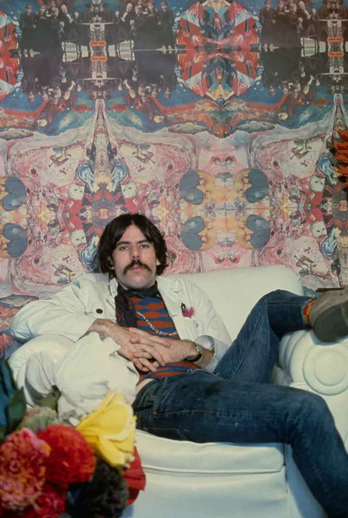 Painter Peter Max poses for a photo while lounging on a white couch in front of a colorful, psychedelic mural.