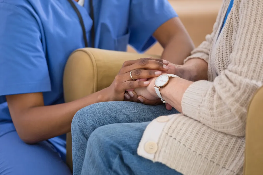 A caregiver in blue scrubs holding the hands of a senior woman in a beige cardigan and jeans.