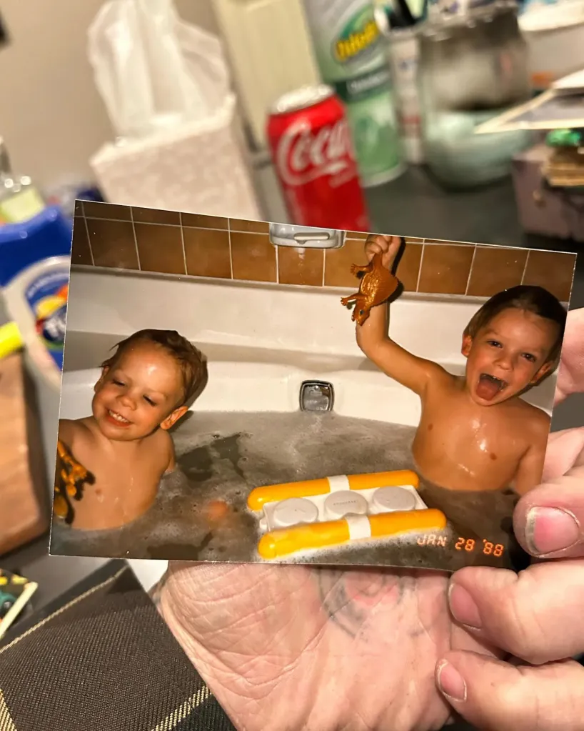 A person holding a faded photo of two young boys bathing together with bath toys.