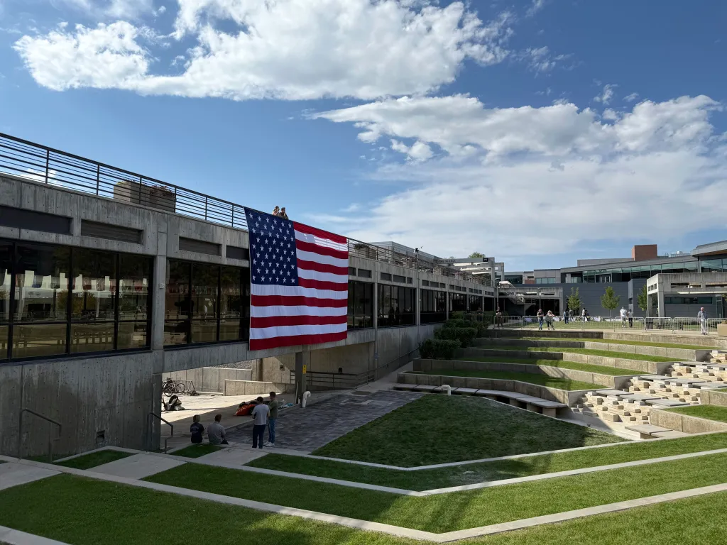 An American flag hangs from a concrete building at Utah Valley University as people stand below and around it.