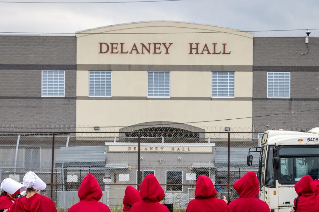 People protesting outside Delaney Hall, an Immigration and Customs Enforcement (ICE) detention center, wearing red cloaks and white bonnets.