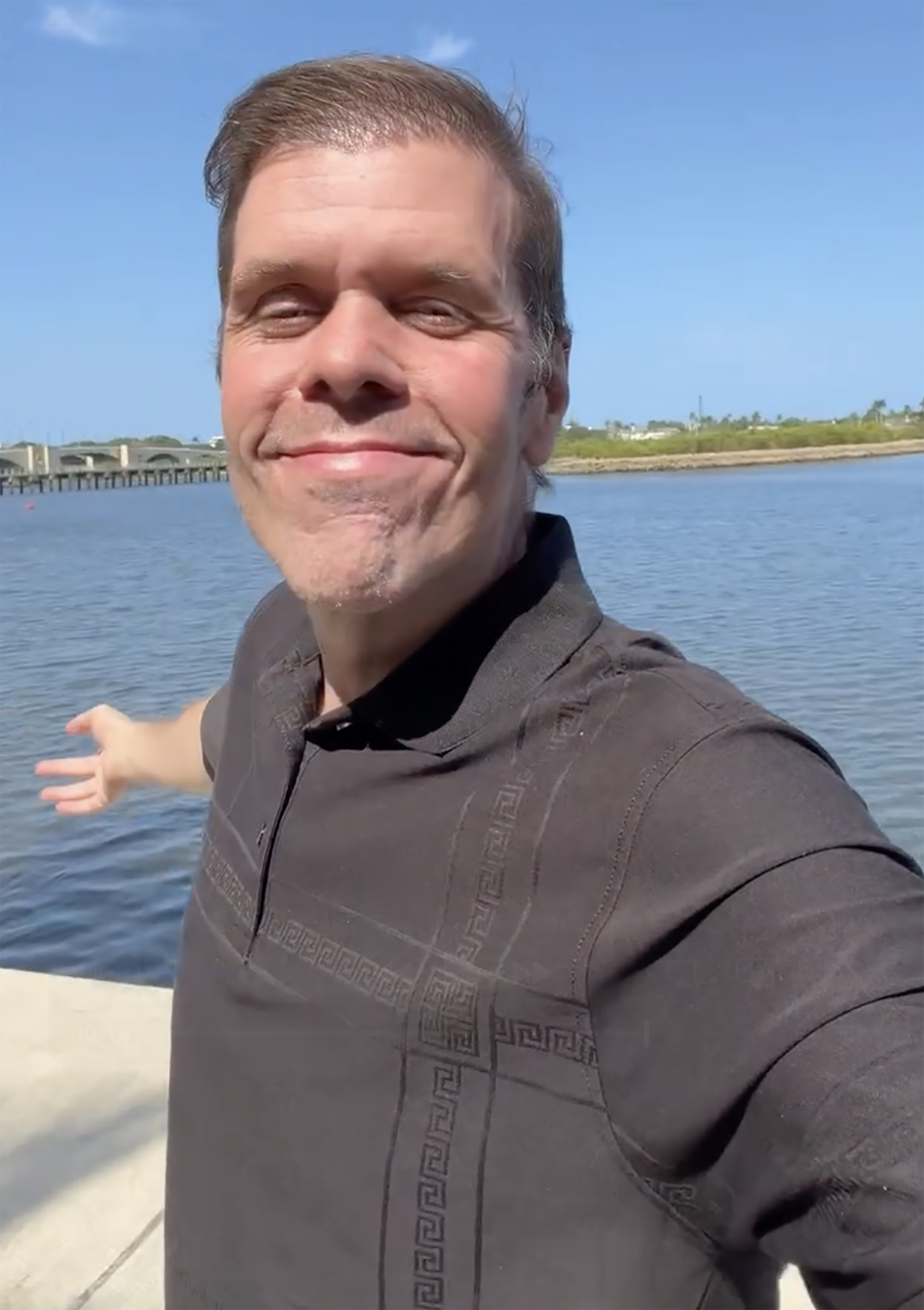 Perez Hilton smiling at the camera outdoors, gesturing to the water behind him.