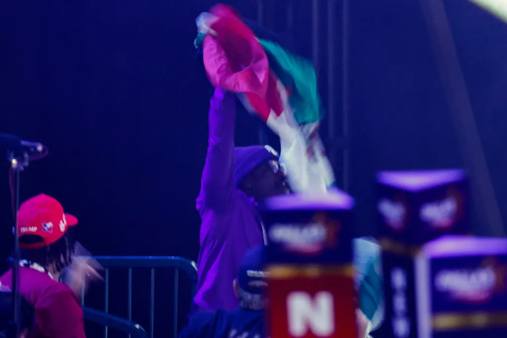 A person in a purple beanie and jacket waves a Mexican flag, while another person in a red 