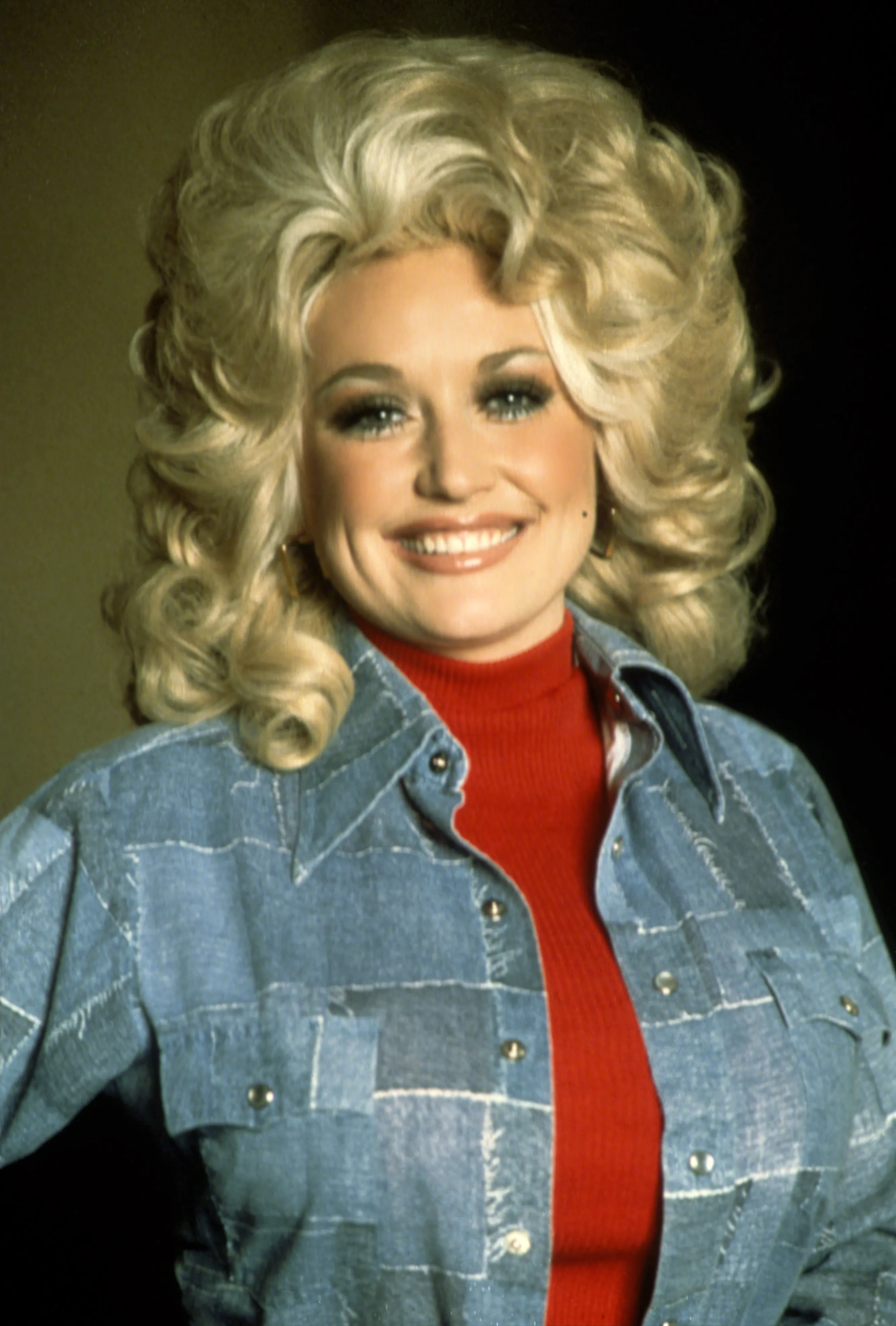 Dolly Parton wearing a red turtleneck under a light blue patched denim shirt.