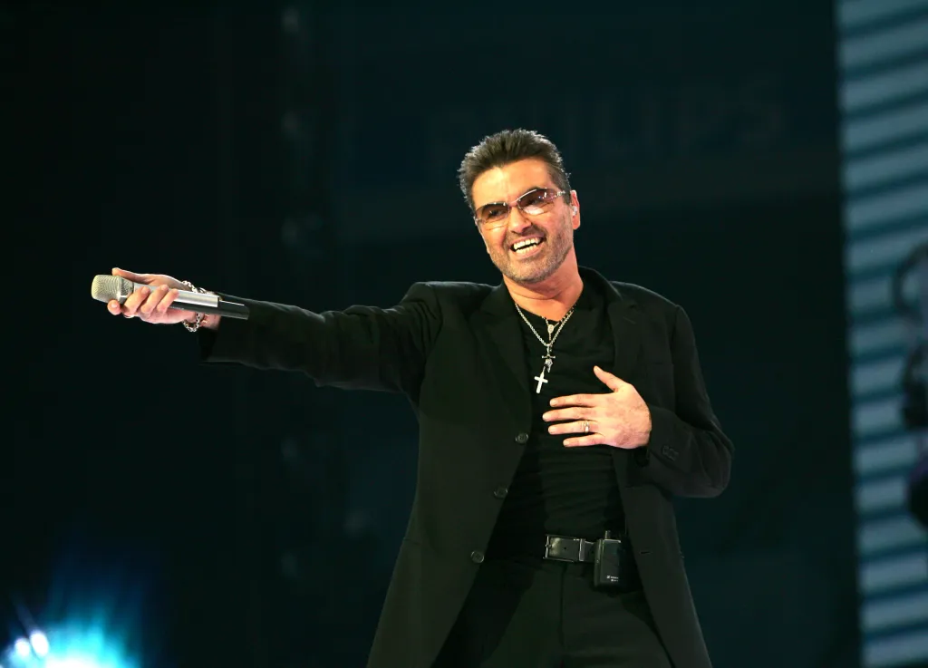 George Michael singing on stage, wearing a black blazer, black t-shirt, and cross necklace, holding a microphone.