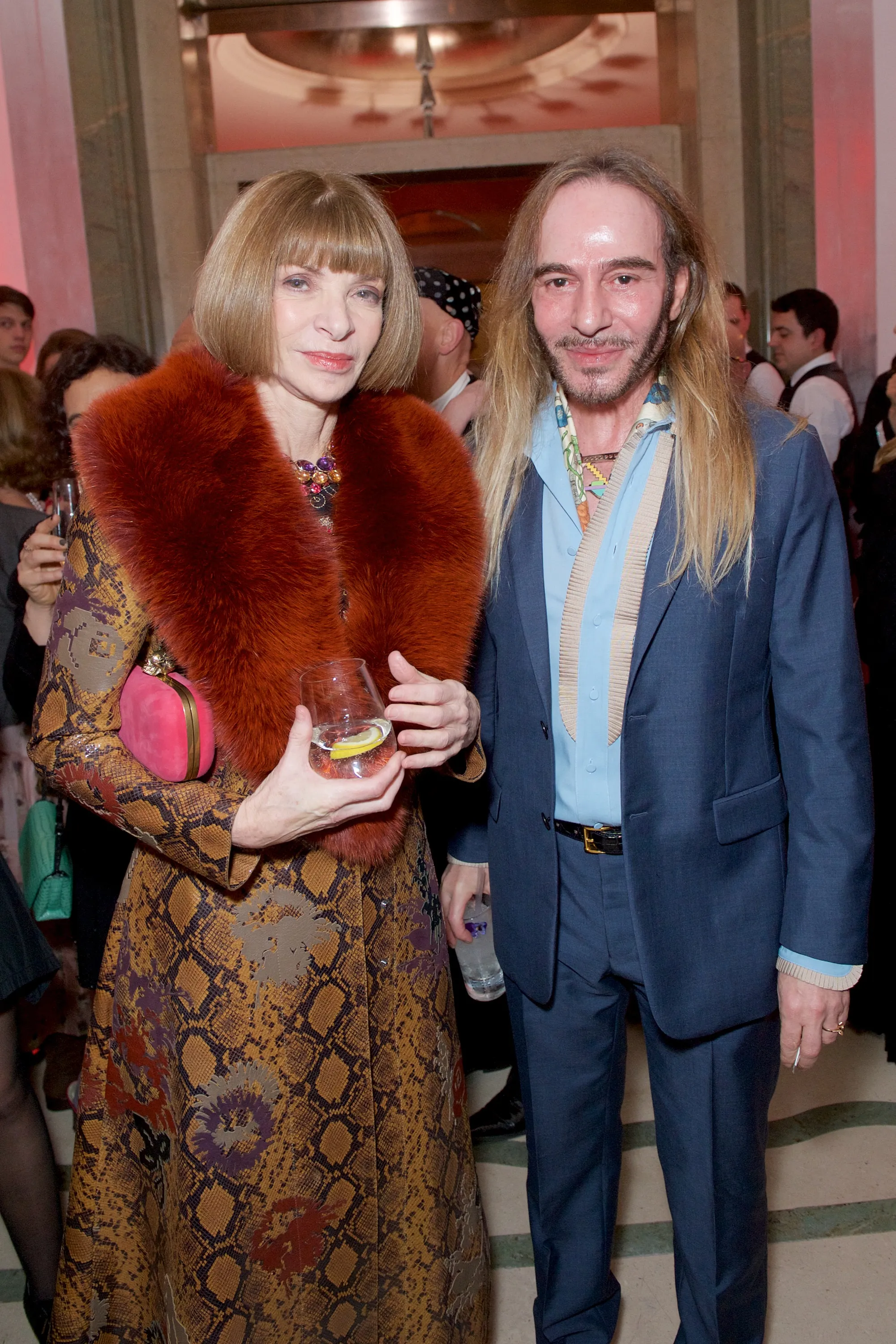 Anna Wintour in a snake-print coat with a fur collar stands next to John Galliano in a blue suit and a patterned scarf.