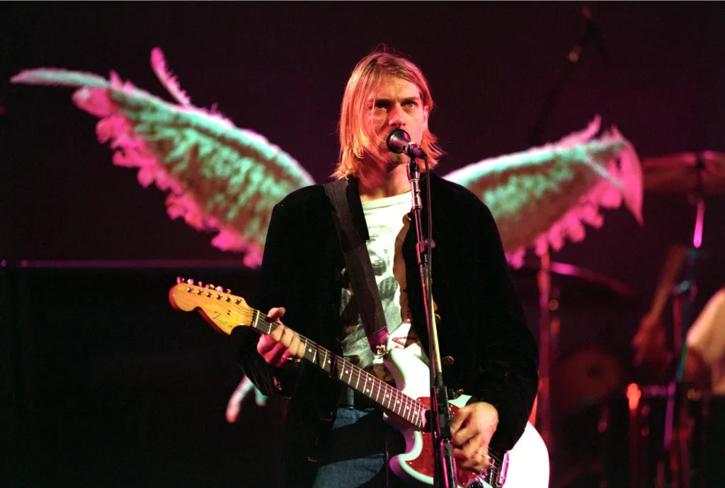 Kurt Cobain playing guitar and singing into a microphone on stage, with large angel wings projected behind him.