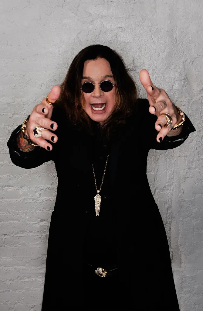 Ozzy Osbourne with dark sunglasses and long black hair, wearing a black shirt, holding out his hands, palms up, with rings on his fingers.