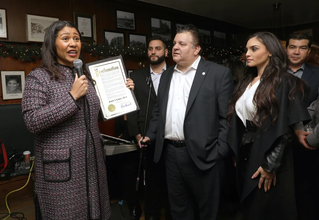 Mayor London Breed announcing the creation of an official John's Grill day, holding a proclamation next to owner John Konstin at John's Grill's 110th anniversary.