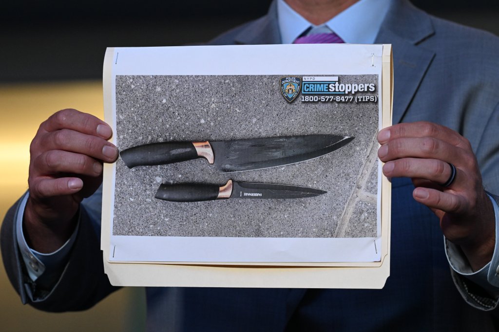 A person in a suit holds up a photo of two knives with the NYPD CrimeStoppers logo.