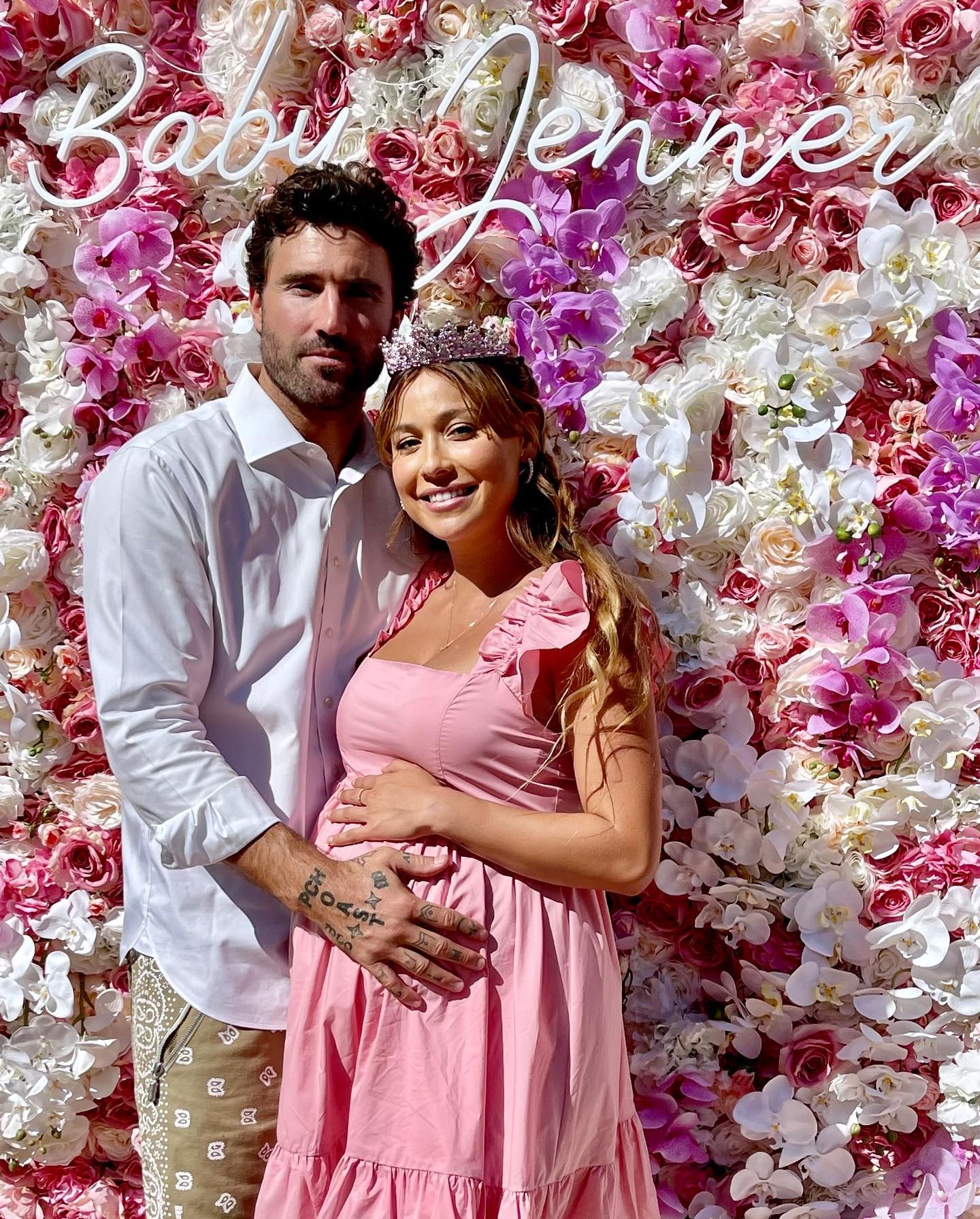 Brody Jenner and Tia Blanco posing together at their baby shower, with Tia wearing a pink dress and a crown, and Brody embracing her in front of a flower wall with a 