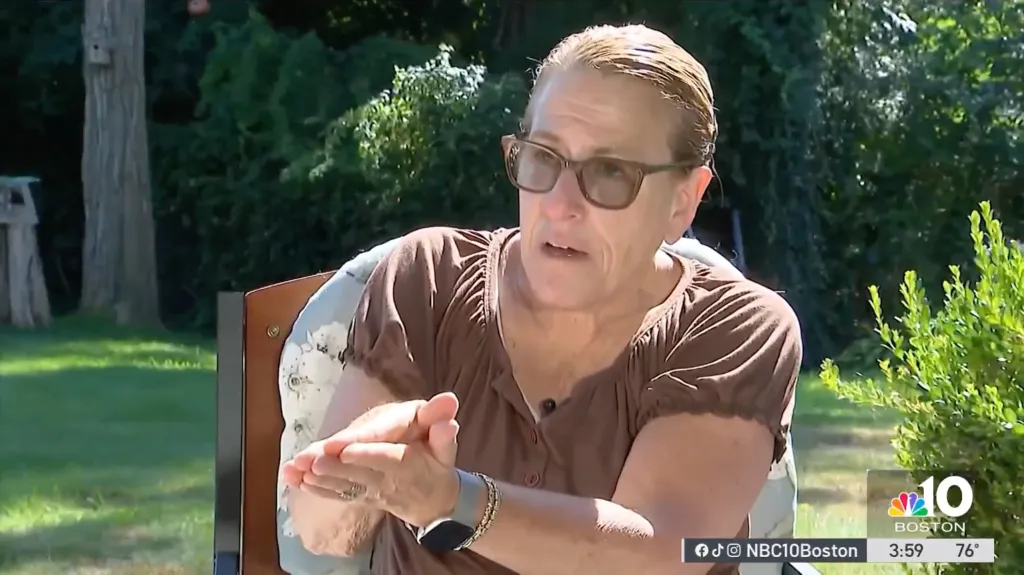 A woman in glasses and a brown shirt talking, gesturing with her hands.