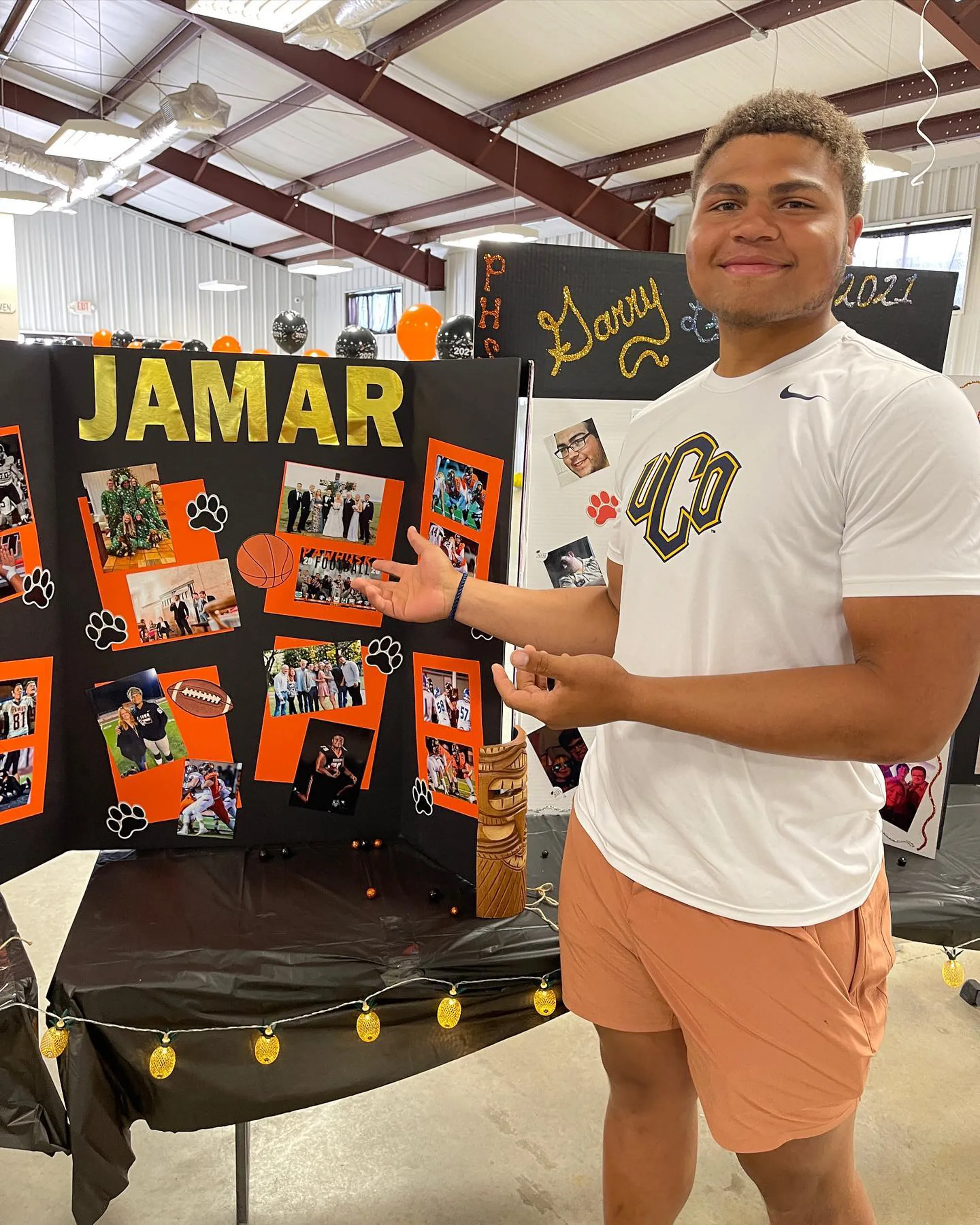 A man in an athletic shirt and shorts stands next to a display board featuring photos, paw prints, and a football and basketball.