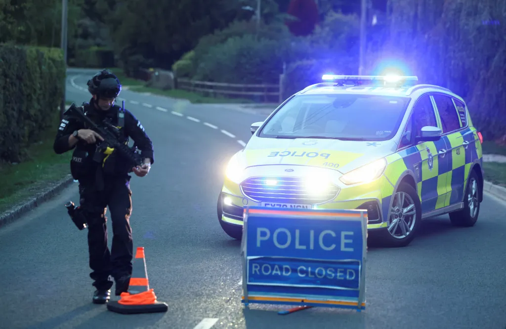 A police officer holding an assault rifle stands by a police car with flashing lights and a 